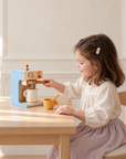 Little girl playing barista with wooden pretend coffee maker at a wooden table.